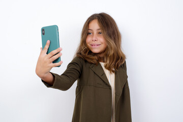 Isolated shot of pleased cheerful Beautiful little girl standing against white background,, makes selfie with mobile phone. People, technology and leisure concept