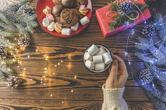 Winter party concept with hot chocolate, hands with cocoa cup with marshmallow on wooden table with christmas decorations, top view layout copy space