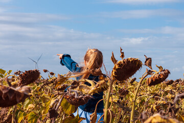 Cute girl in denim jacket smelling showing finger on wind turbines farm on sunflowers field background. Child with long hair on countryside evening sunset landscape with yellow flowers.Farming concept