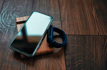 A cell phone (buttonless) and a smart bracelet lie on a wooden table stand. The light from the window hits the screen at an angle.