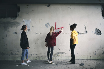 Rear view of group of teenagers girl gang indoors in abandoned building, using spray paint on wall.
