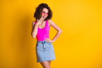 Photo of attractive girlish wavy lady look interested side empty space sly coquettish girlfriend arm near mouth wear pink sun glasses singlet denim skirt isolated yellow shine color background
