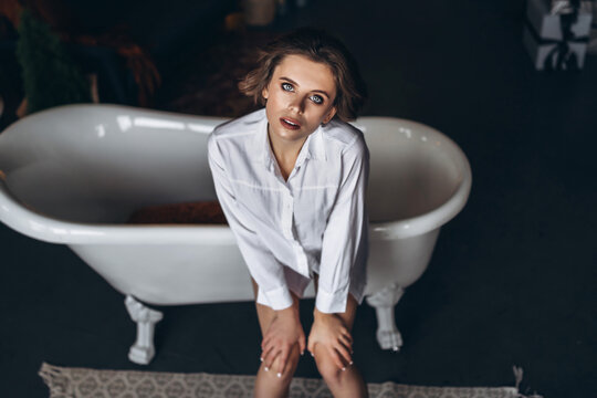 Top view of a young brunette with short hair sitting on the edge of a bath in a white shirt.