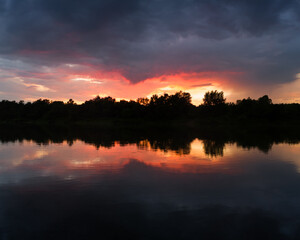 The landscape of the river after the storm during the summer evening at sunset, the reflection in the water, the dark dramatic clouds and the sunshine beyond the horizon in red and orange.