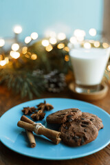 Fototapeta na wymiar Chocolate chip cookies on plate and glass of milk for Santa placed on wooden table with glowing garland 