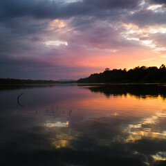 The peaceful landscape of the river and mountain peaks in the distance during a cloudy summer evening, at sunset, the sunlight behind the horizon illuminates the clouds in orange and pink.