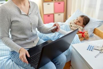 family, health and people concept - mother with laptop computer and thermometer measuring temperature of sick daughter lying in bed at home