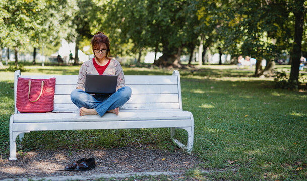 Mature woman with laptop outdoors in city or town park, working.