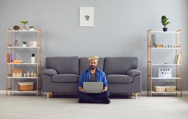Young smiling man sitting on floor with laptop on knees and working online from home