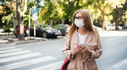 Senior woman with smartphone crossing street outdoors in city or town, coronavirus concept.