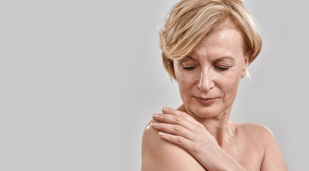 Close up portrait of beautiful middle aged woman looking down, touching her shoulder, posing isolated against grey background. Beauty, skincare concept