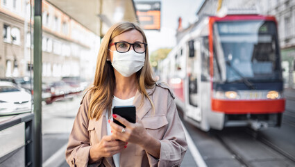 Senior woman with smartphone on bus stop outdoors in city or town, coronavirus concept.