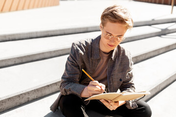 leisure, education and people concept - young man or teenage boy in glasses with notebook, diary or sketchbook writing or drawing in city