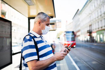 Mature man with smartphone on bus stop outdoors in city or town, coronavirus concept.