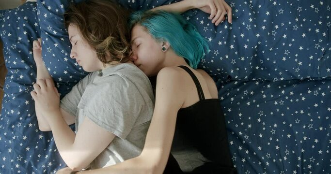 Top view of young woman with dyed hair hugging girlfriend from behind while sleeping on soft bed at home together