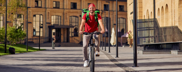 Delivering food to customers. Full length of a young cheerful caucasian male courier with thermo bag riding bicycle along sunny street