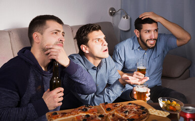 friends in suspense watching tv together at home, drinking beer with pizza