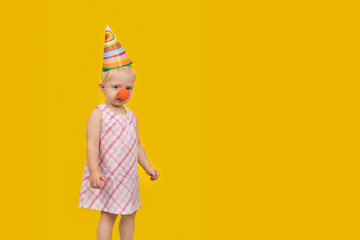 Toddler girl wear party hat and red clown nose on yellow background. Childrens holiday. Copy space