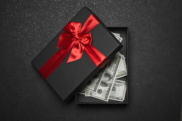 An open black gift box with a red ribbon and a large bow with money inside. Granite background.