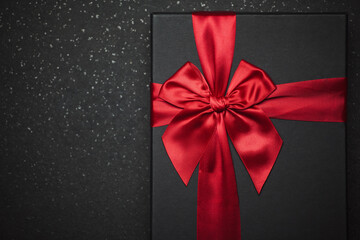 Black gift box with a red ribbon and a large bow on a granite dark background. Close-up photo. Place for the inscription.