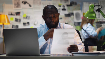 Portrait of afro-american policeman studying identikit of criminal sitting in office - obrazy, fototapety, plakaty