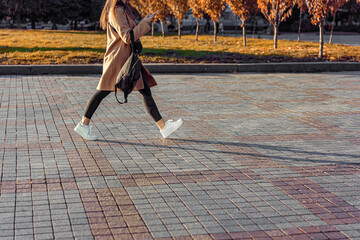 A young woman walks quickly along the wide sidewalk. Beige coat, black pants, white sneakers. Autumn, sunny weather