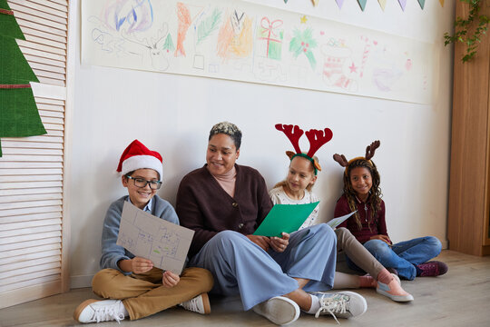 Full length portrait of female teacher sitting on floor with multi-ethnic group of kids holding pictures while enjoying art class on Christmas, copy space