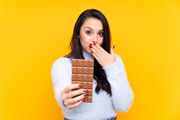 Young Colombian girl over isolated yellow background taking a chocolate tablet and surprised
