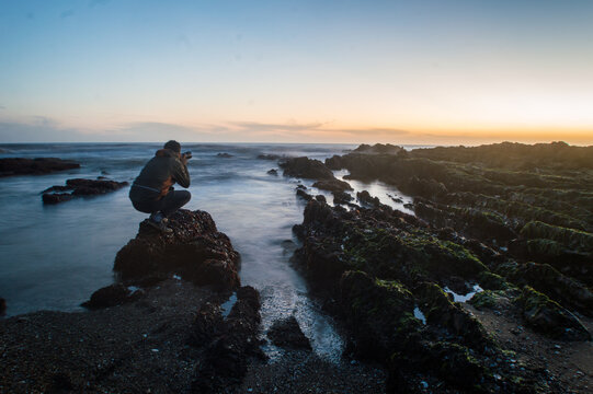 fot&oacute;grafo agachado sobre una roca tomando una fotograf&iacute;a del atardecer