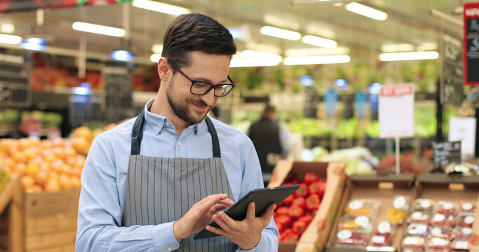 Close up portrait of happy Caucasian male worker in glasses standing in supermarket and typing on tablet. Young joyful man food store assistant at work tapping on device indoors. Retail concept