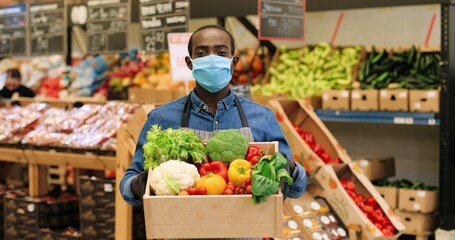 Portrait of joyful male food store manager in mask and gloves standing in supermarket and holding box with fresh vegetables, looking at camera and smiling. African American mam working at grocery.