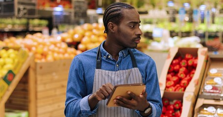 Close up portrait of busy African American male worker standing in supermarket and typing on tablet while doing inventory. Young man food store assistant at work tapping on device. Retail concept