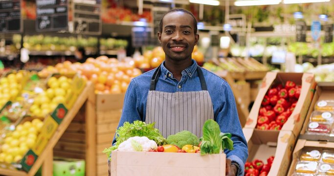 Close up portrait of happy male employee with box with fresh organic vegetables, looking at camera in good mood. Young African American cheerful man manager work at food shop. Business concept