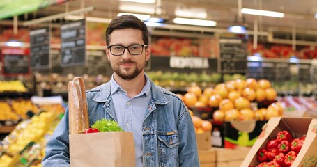 Close up portrait of Caucasian cheerful man in glasses standing in grocery shop with packet with food and vegetables. Happy young handsome guy on shopping at supermarket. Retail concept