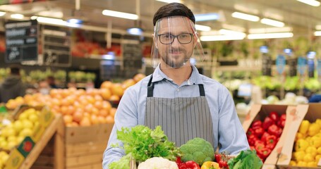 Close up portrait of happy smiling Caucasian male food store worker in face shield and gloves holding in hands box with fresh vegetables and looking at camera. Young guy seller standing in supermarket