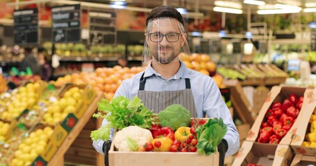 Close up portrait of happy smiling Caucasian male food store worker in face shield and gloves holding in hands box with fresh vegetables and looking at camera. Young guy seller standing in supermarket