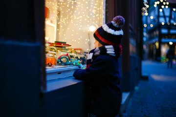 Cute toddler boy looking on car toys in a window on Christmas time season. Fascinated child in winter clothes dreaming and wishing. Window decorated with xmas gifts. Snow falling down, snowfall.