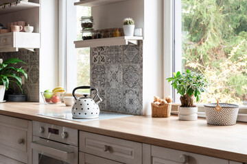 Cozy white kitchen with wooden furniture and counter with design tiles and window with plant....