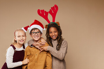Waist up portrait of three children wearing Santa hats smiling at camera while standing against beige background, copy space