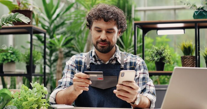 Close up portrait of handsome cheerful male florist buying online on smartphone and paying with credit card at work in floral store. Young happy businessman in apron shopping on internet on cellphone