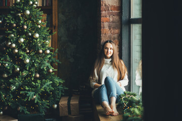 A beauty young girl sitting on the windowsill near christmas tree at home. christmas morning