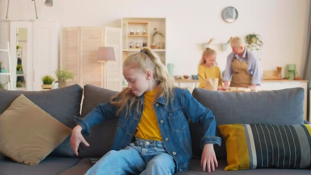 Medium long of blond-haired Caucasian school girl jumping on couch, taking TV remote, pushing buttons on it and sitting. Grandma and sister cooking in kitchen in background