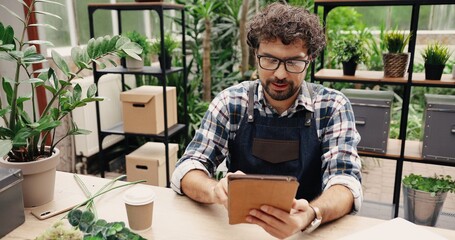 Close up of Caucasian man entrepreneur in glasses sitting in own flower shop and typing on tablet indoor. Young male florist in floral store working and texting on device. Floral concept