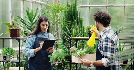 Portrait of happy young Caucasian couple working in own flower shop indoor Joyful female entrepreneur tapping on tablet while male florist watering plants with sprayer in floral center Family business