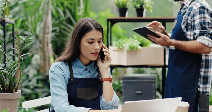 Beautiful Busy Woman Entrepreneur In Apron Sitting In Own Floral Store And Talking On Smartphone With Client. Caucasian Male Florist Typing On Tablet While Boss Speaking On Cellphone. Flower Shop