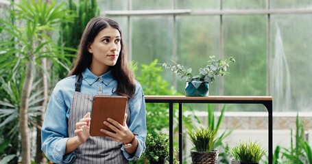 Portrait of Caucasian happy young pretty woman florist in apron typing on tablet at floral store. Beautiful female owner of flower shop tapping on device while working indoor. Working concept