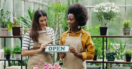 Happy mixed-races women florists in aprons standing in own floral shop and smiling to camera with Open sign in hands. Caucasian and African American females reopening flower store. Portrait concept