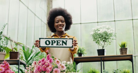 Portrait of African American joyful woman florist in apron standing at flower shop and smiling to camera with Open sign in hands Happy female entrepreneur running own floral business Reopening concept