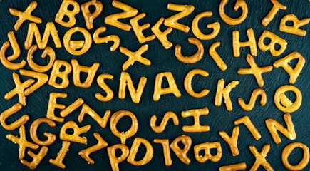Snack text among salty sticks letters, alphabet on black stone background. Junk food, unhealthy diet concept.