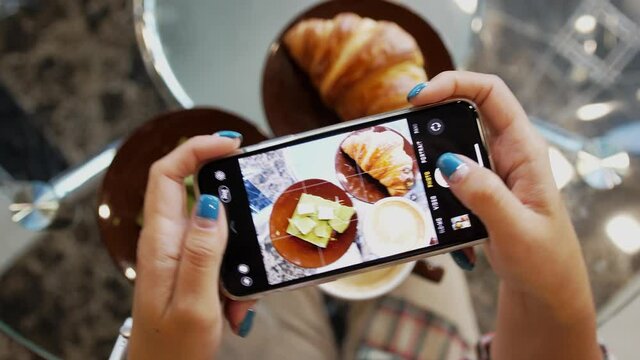 Close up of woman holds modern smartphone and taking pictures of her morning breakfast coffee, green chocolate and tasty croissant buns to share photos on social media resources